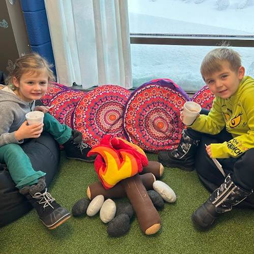 A boy and girl sip cocoa around a pretend bonfire made of pillows